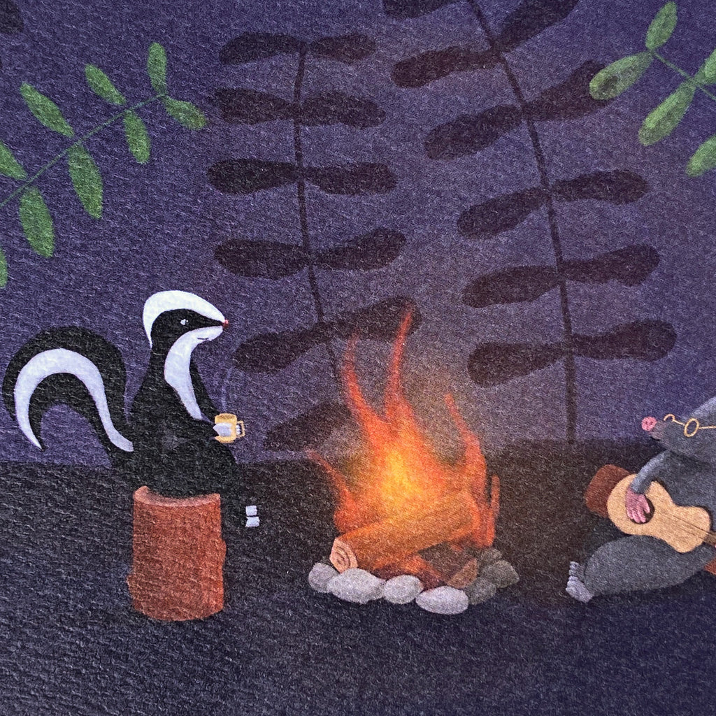 A close up of a greeting card with a whimsical illustrated scene showing a black-and-white skunk sitting on a brown log by a glowing campfire, with a gray mole playing guitar at the right. The background is a dark purple textured field with leafy silhouettes.