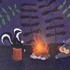 A close up of a greeting card with a whimsical illustrated scene showing a black-and-white skunk sitting on a brown log by a glowing campfire, with a gray mole playing guitar at the right. The background is a dark purple textured field with leafy silhouettes.