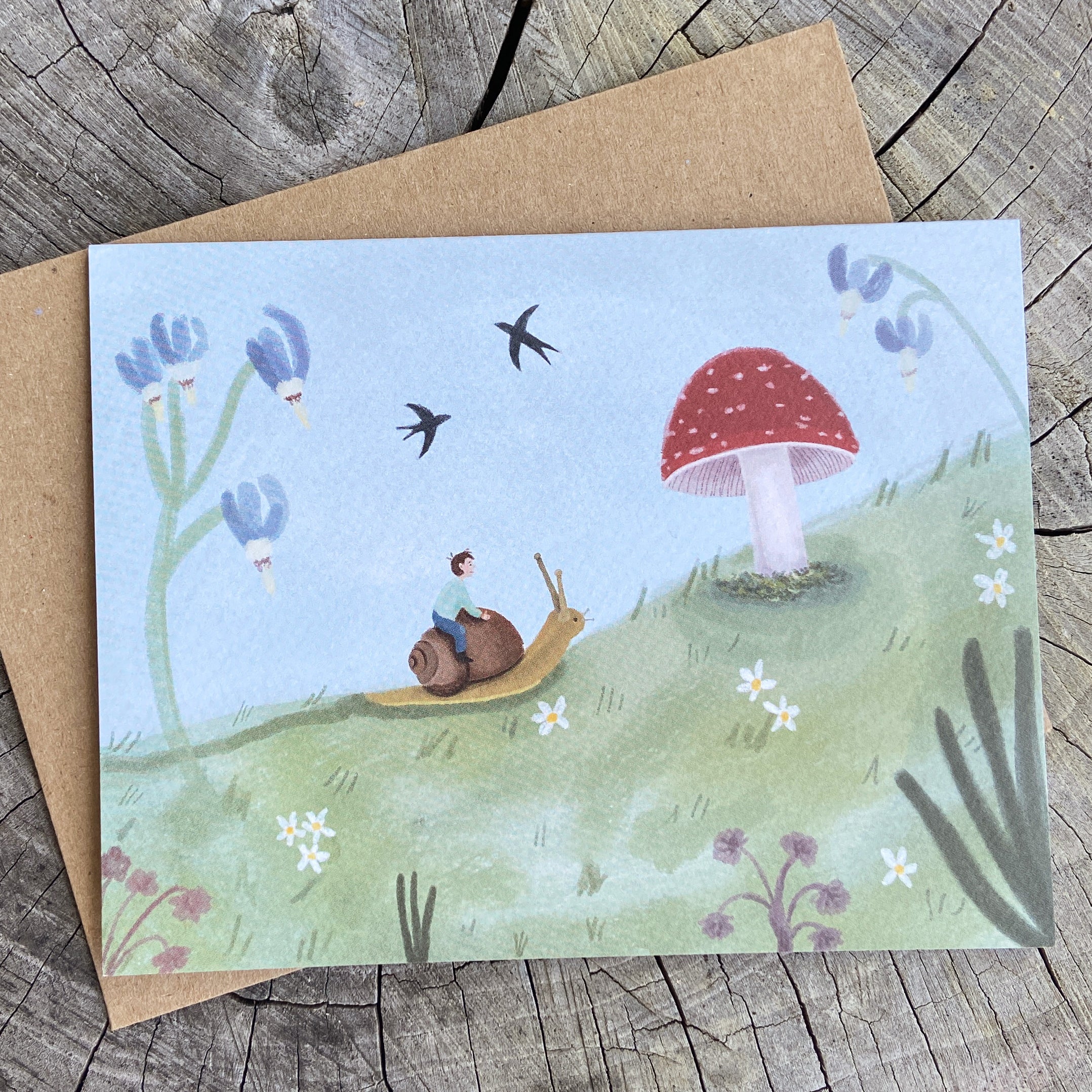 Whimsical greeting card with illustration of a child riding a snail near a mushroom, on a wooden surface