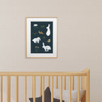 Framed artwork with white rabbits on a dark  background above a wooden baby crib.