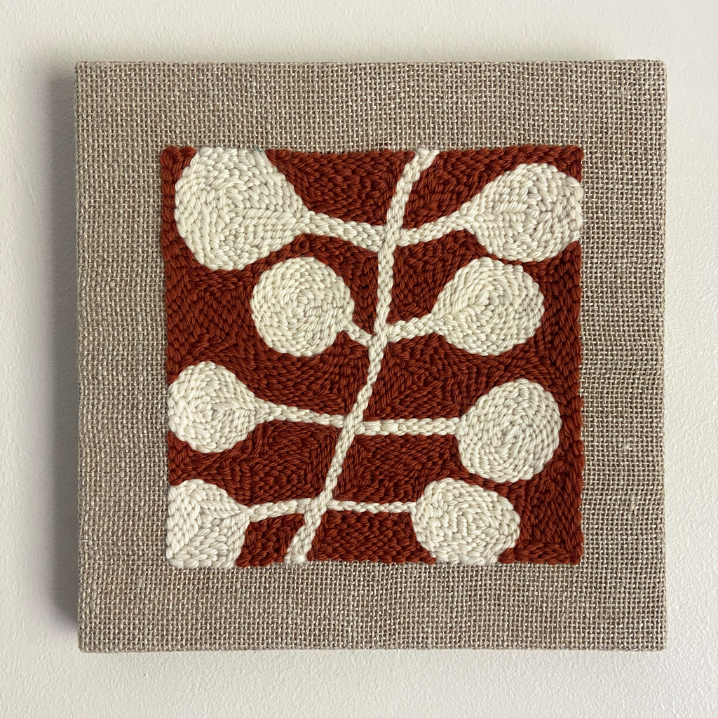 A square woven wall hanging or tapestry with a textured beige linen border and a central rust red square featuring an abstract cream leaf or vine pattern.