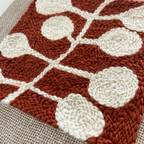 A close-up of a handwoven textile with a terracotta rust and off-white geometric pattern, laid flat on a beige woven surface.