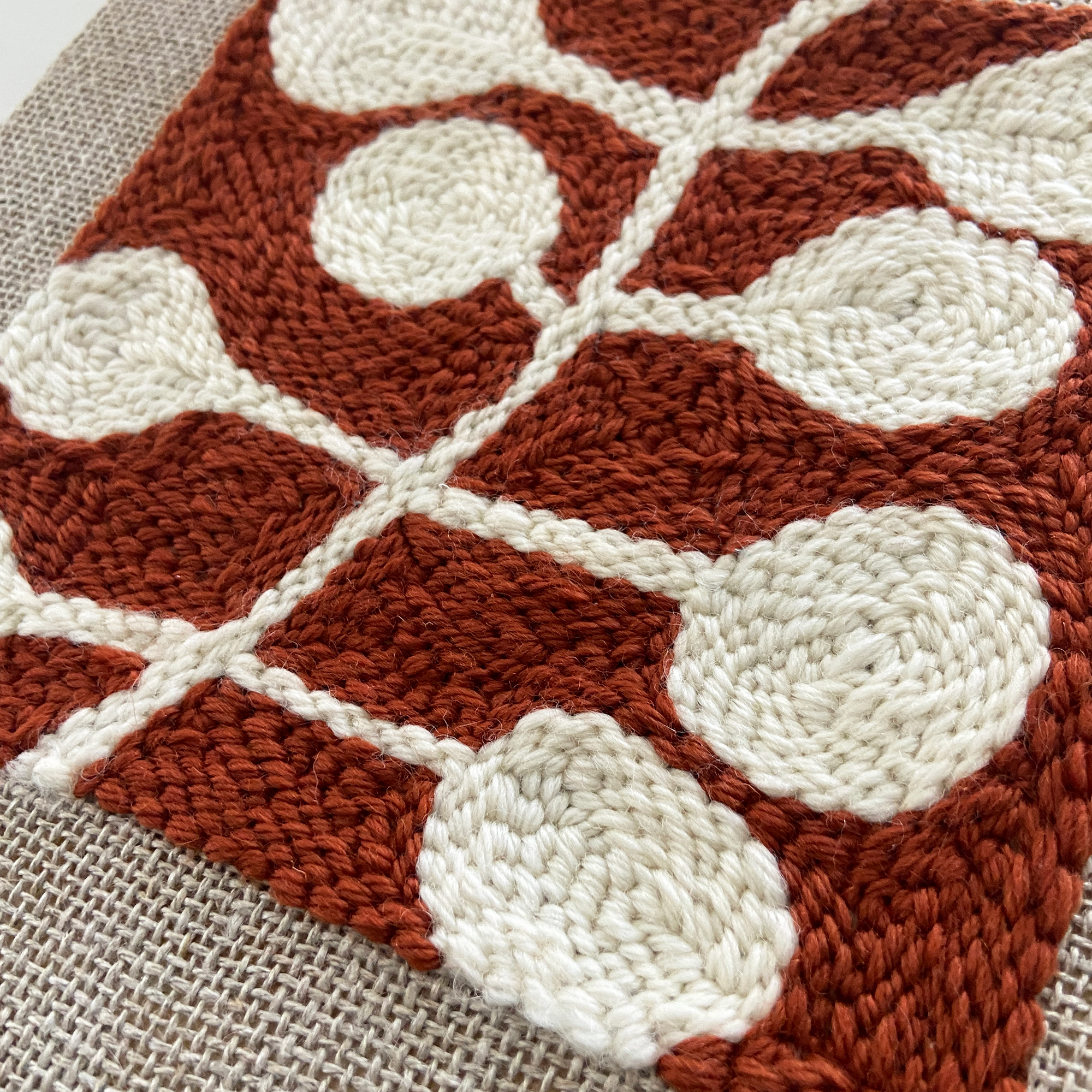 A close-up of a handwoven textile with a terracotta rust and off-white geometric pattern, laid flat on a beige woven surface.