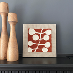 A square framed textile artwork with a red-orange background and white abstract leaf-like motifs, displayed on a dark mantel beside two tall wooden candlesticks against a gray wall.