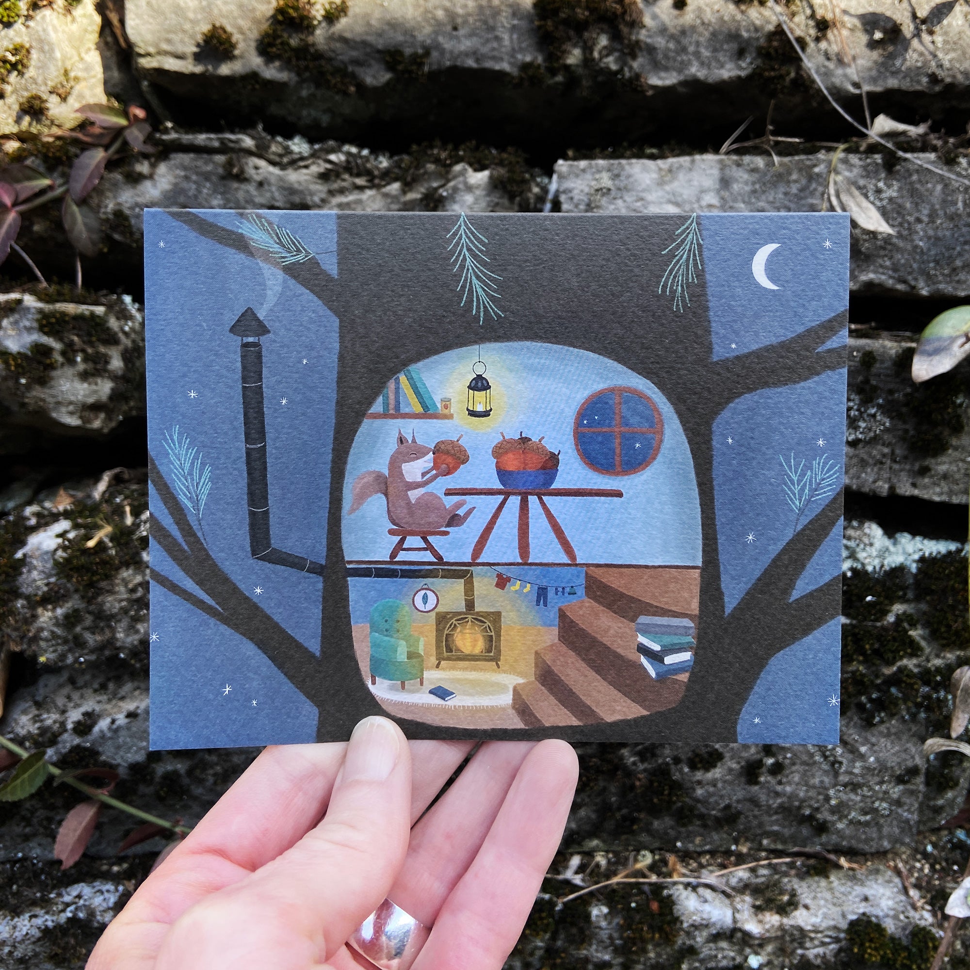 Illustrated card of a cozy treehouse scene with a squirrel eating acorns, held over a natural background