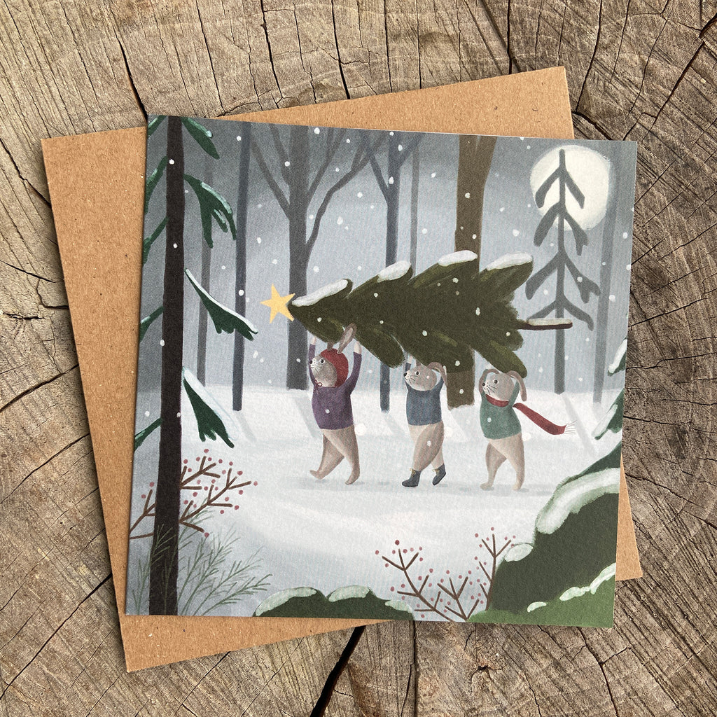 Holiday greeting card with illustration of bunnies carrying a Christmas tree in a snowy forest.
