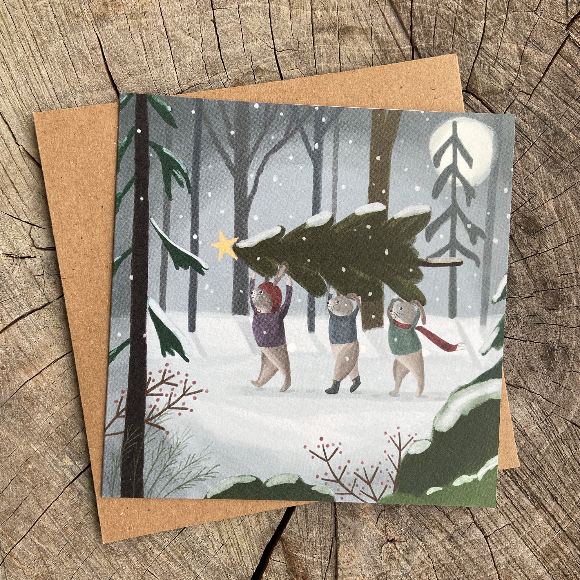 Holiday greeting card with illustration of bunnies carrying a Christmas tree in a snowy forest.