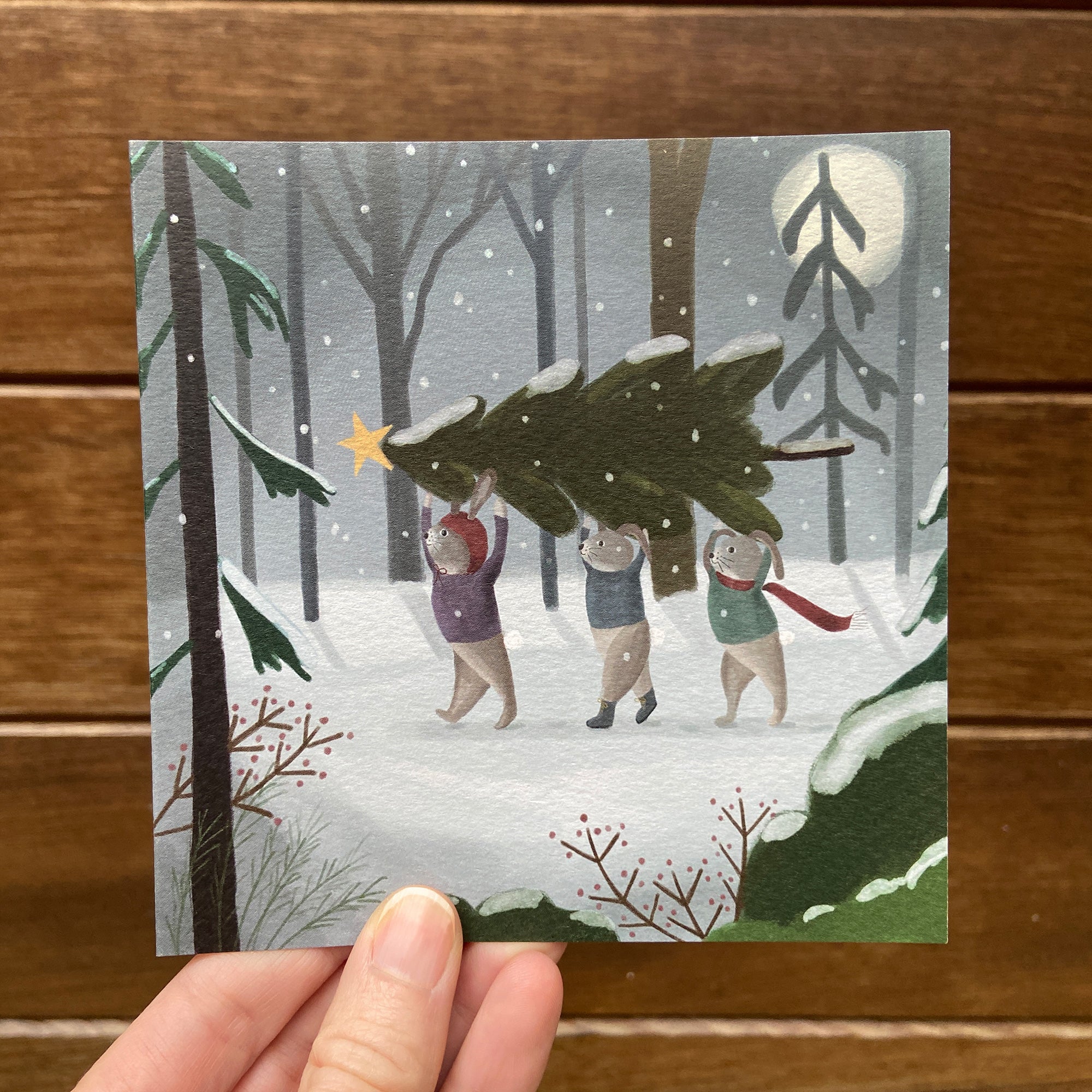 Illustration of rabbits carrying a Christmas tree in a snowy forest, held against a wooden background.