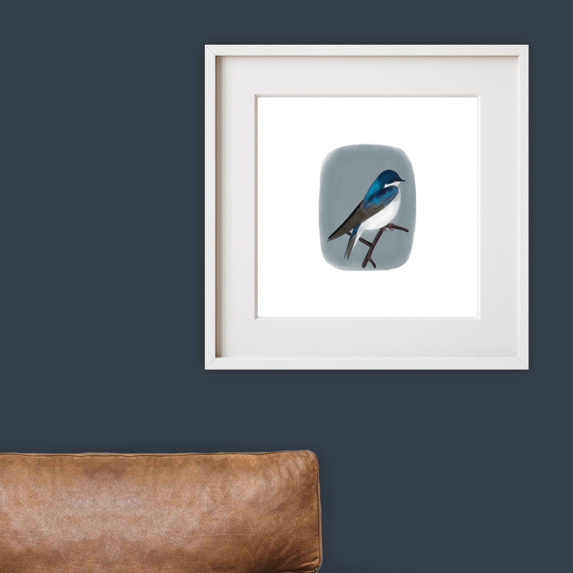 Framed artwork of a tree swallow on a gray background above a brown leather couch.