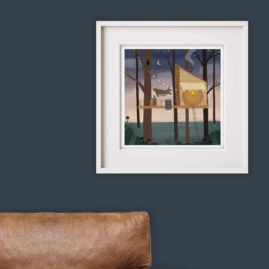 Framed artwork of a treehouse in a forest with a woodland bear on a dark blue wall.