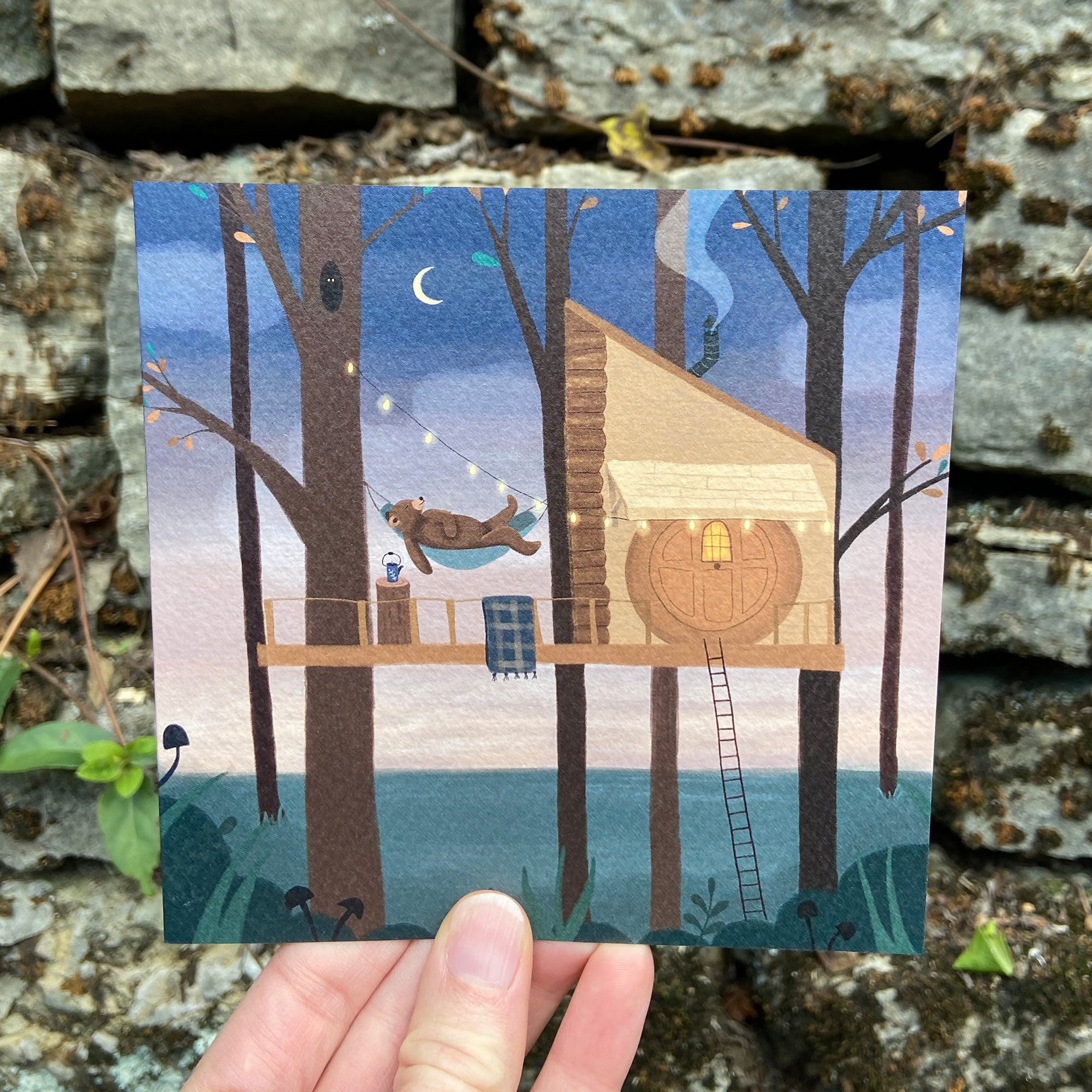 Illustrated greeting card of a treehouse and bear relaxing in the woods.