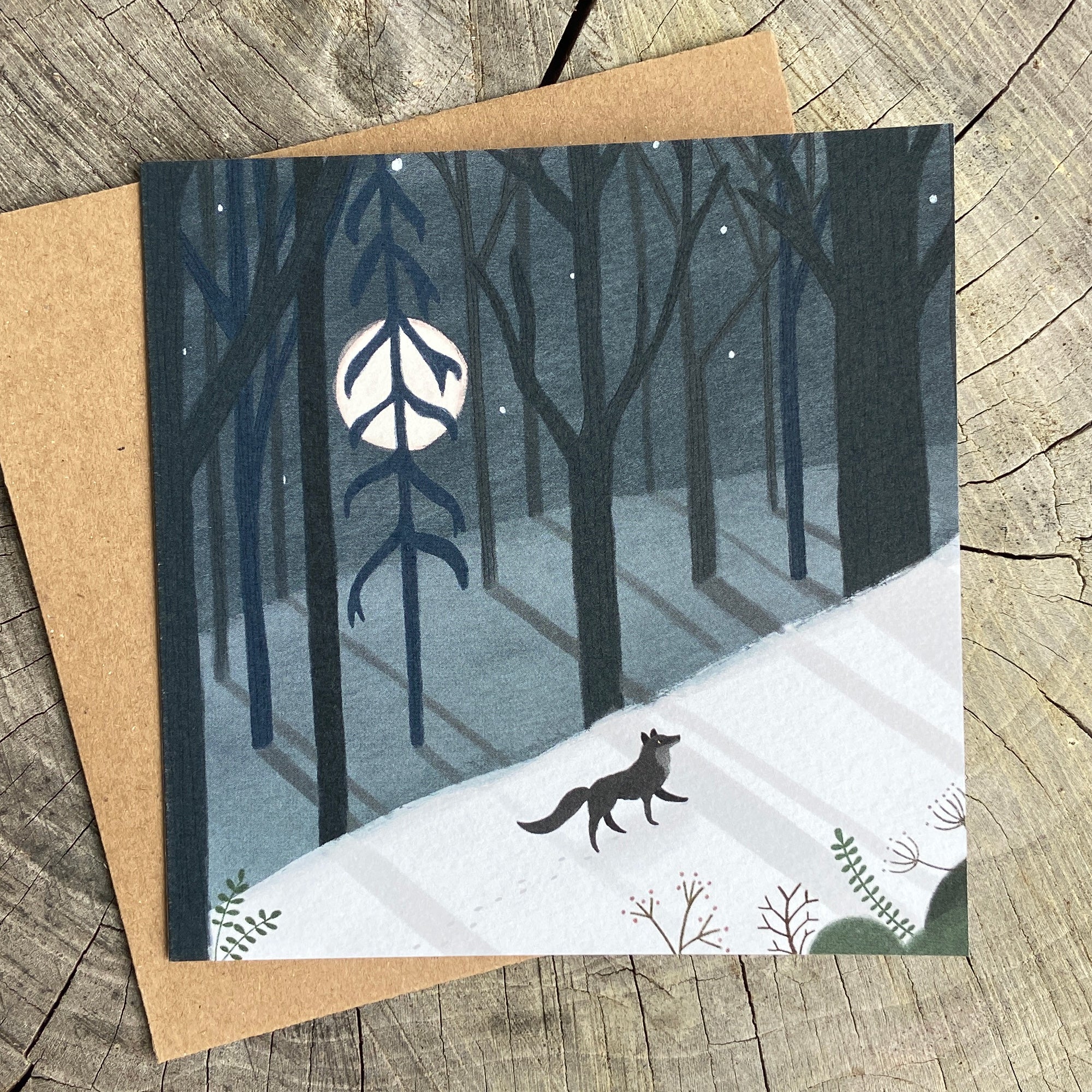 Greeting card with a winter forest design featuring a wolf and a full moon, on a wooden surface.